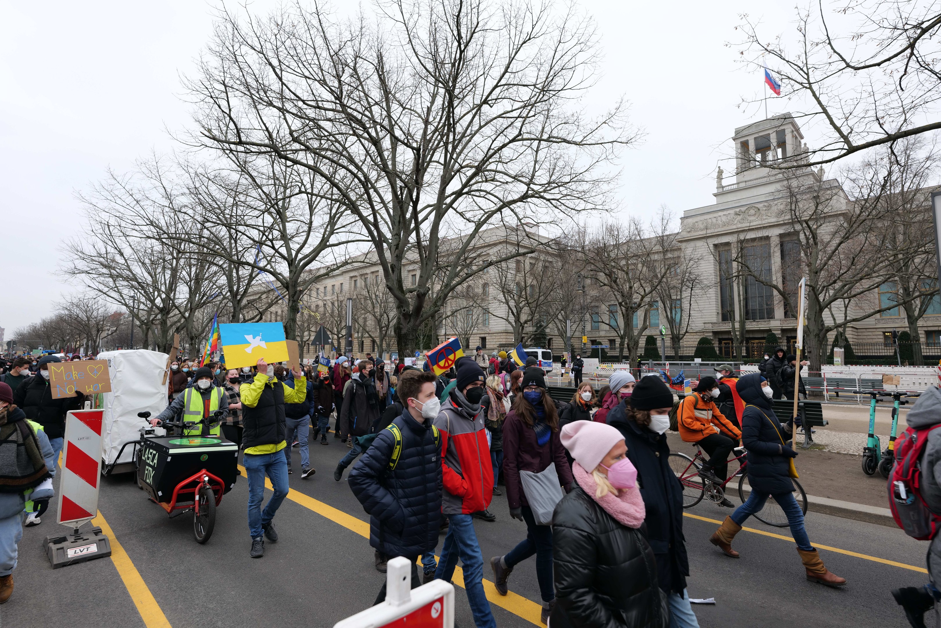 Eine große Gruppe von Menschen marschiert auf einer Straße in Washington, D.C. und hölt Plakate und Schilder, während einige Fahrráder fahren, unter einem klaren blauen Himmel.