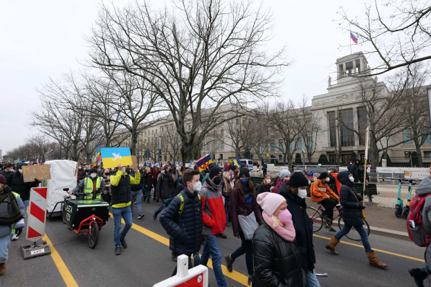 Eine große Gruppe von Menschen marschiert auf einer Straße in Washington, D.C. und hölt Plakate und Schilder, während einige Fahrráder fahren, unter einem klaren blauen Himmel.