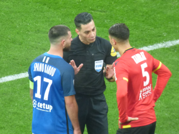 Schiedsrichter in schwarzer Uniform, der mit zwei Männern auf einem Fußballfeld spricht, einer in einem blauen und roten T-Shirt mit Text und Brille, der andere ebenfalls mit Brille.
