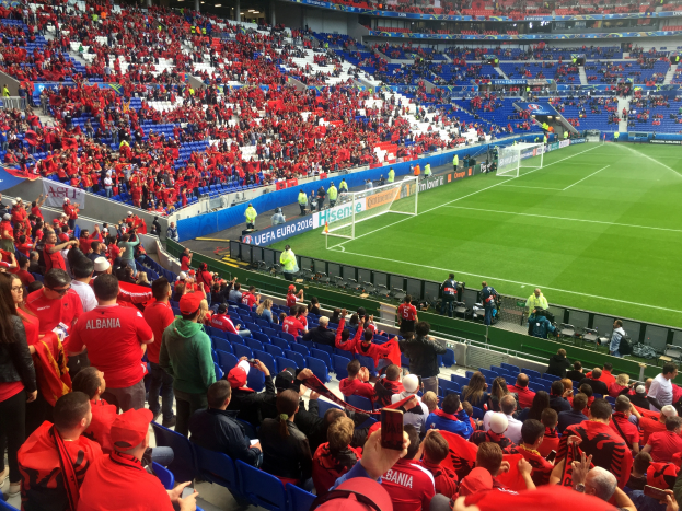 Ein volles Stadion mit Zuschauern, einige sitzen und einige stehen, die ein Fußballspiel beobachten, mit Bannern, einem Torpfosten und Deckenleuchten.