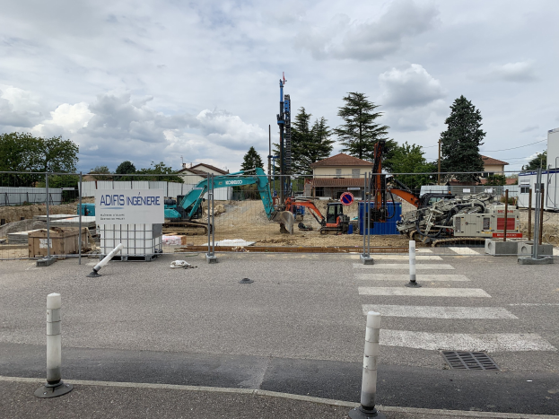 Baustelle mit verschiedenen Geräten wie Pfählen, einem Zaun, einem Schild, Fahrzeugen, Bäumen, Häusern und einem bewölkten Himmel.