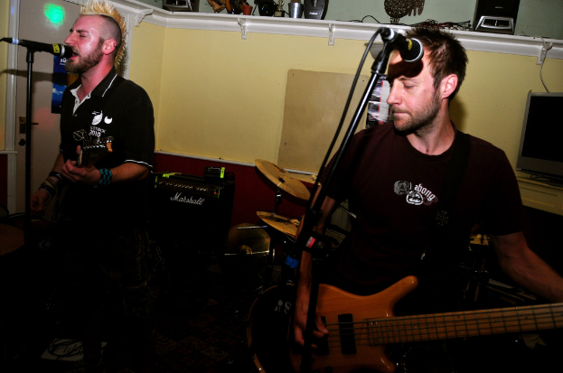 Zwei Personen stehen und spielen Gitarren, eine singt in ein Mikrofon, mit Trommeln dahinter und einer Tafel an einem Schrank im Hintergrund.