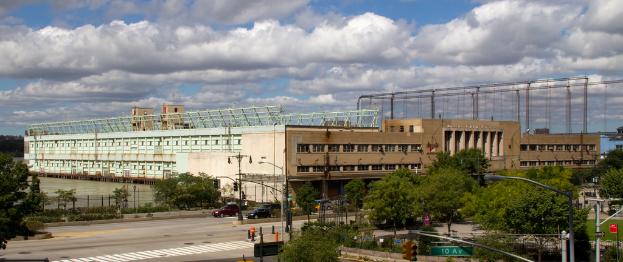 Eine Straße mit Fahrzeugen, Bäumen und Straßenlaternen entlang einer Wasserfläche, mit einem großen Gebäude im Hintergrund.