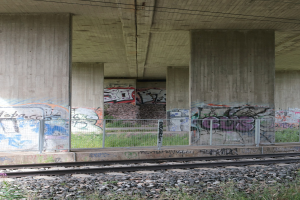 Zuggleis mit Graffiti an der Seite, umgeben von Steinen, Gras, einem Zaun, Säulen, Drähten und einem Dach.