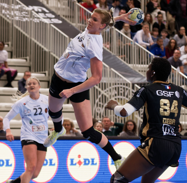 Eine Gruppe von Frauen spielt Handball auf einem Platz mit einem Ball in der Luft, Zuschauern auf Treppen und einem "Handball-Weltmeisterschaft 2019 - Tag 2"-Schild im Hintergrund.