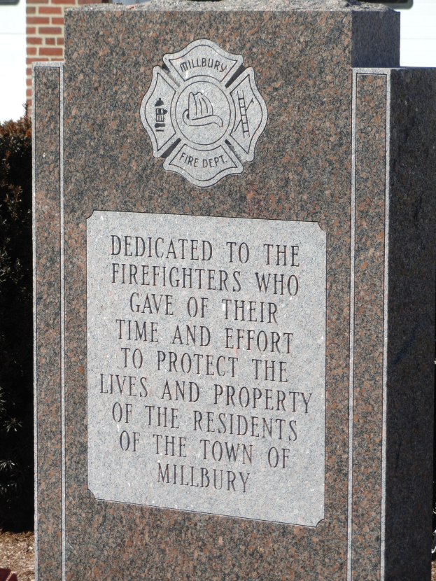 Gedenkstein mit einer Gedenktafel für Feuerwehrleute in Millbury, aufgestellt vor einem Backsteingebäude mit umgebender Vegetation.