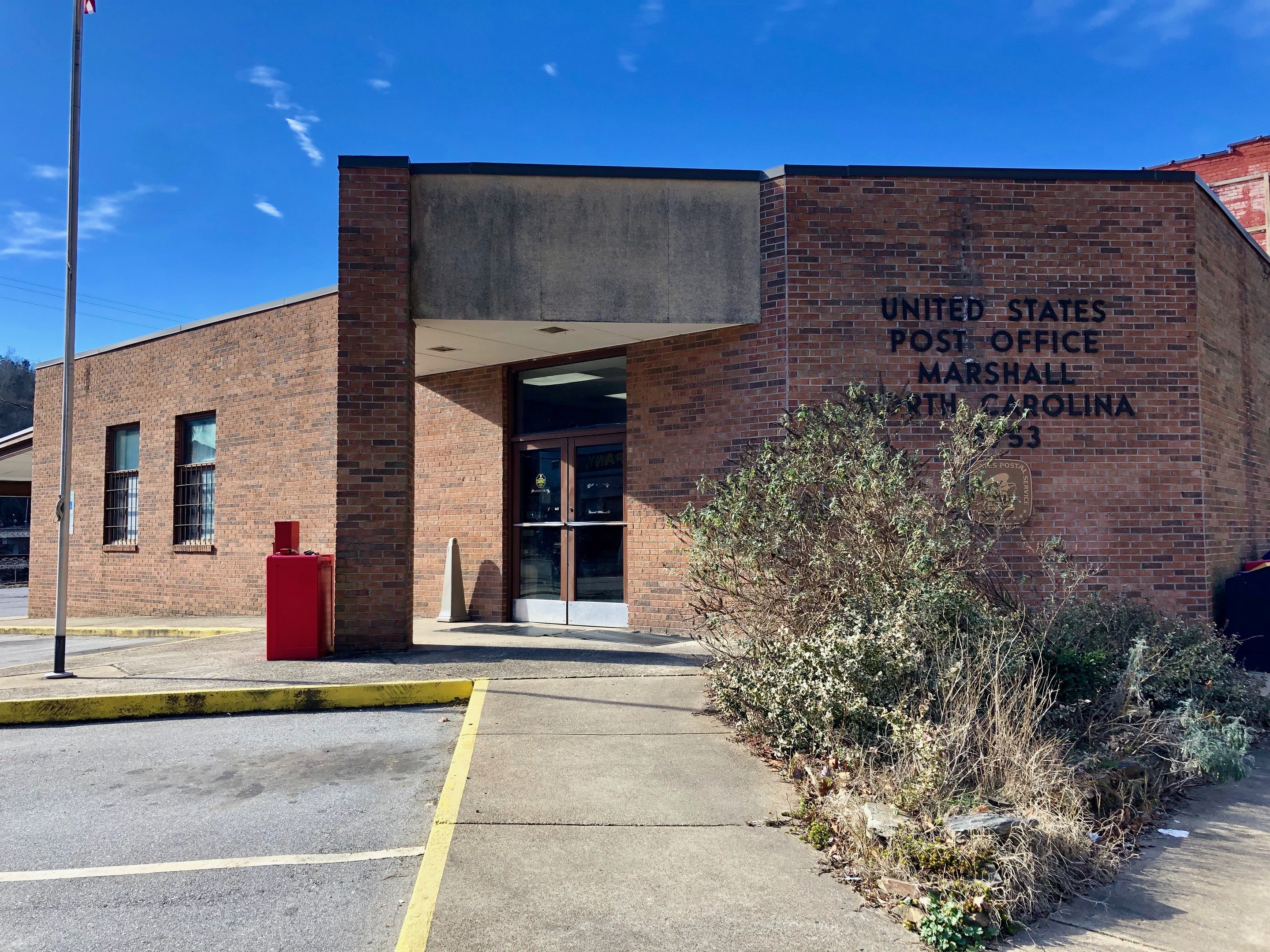 Außenansicht der Vereinigten Staaten Post Office in Marshall, North Carolina, mit einem Backsteingebäude mit Glastüren und -fenstern, einer Flagge an einem Mast mit einer amerikanischen Flagge, einem roten Briefkasten, Pflanzen, einem gepflasterten Weg, einem geparkten Auto, umliegenden Bäumen und einem bewölkten Himmel.