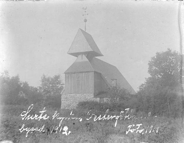 Schwarzes und weißes Foto einer Kirche mit einem Turm, umgeben von Bäumen und Pflanzen unter einem bewölktem Himmel, beschriftet mit "Kirche des Heiligen Grabes" unten.
