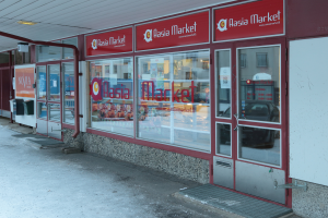 Ein Geschäft mit einer Tür, die mit "Asia Market" beschriftet ist, Glaswänden und -türen, Deckenleuchten und Schildern; links sind Mülltonnen und andere Gegenstände zu sehen.