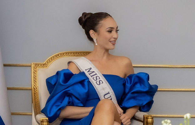 Frau in einem blauen Kleid mit einem "Miss Universe 2019" Band, sitzend auf einem Stuhl umgeben von Blumen mit einem Lächeln im Gesicht.