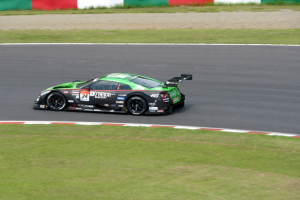 Ein rotes Sportauto mit Text und Zahlen auf seiner Karosserie fährt auf einer Rennstrecke, die von Gras gesäumt ist, mit einer Wand im Hintergrund während der 24 Stunden von Le Mans.