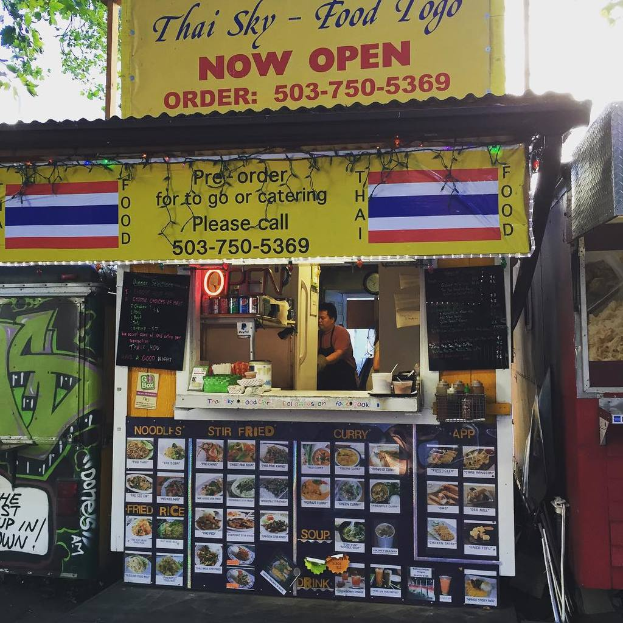 Eine Straßenansicht mit Schaufenstern mit verschiedenen Schildern, Speisekarten und Werbetafeln sowie Regalen mit Getränkedosen, Gewürzen und Gewürzbehältern aus Plastik und einer Uhr in der Szene.