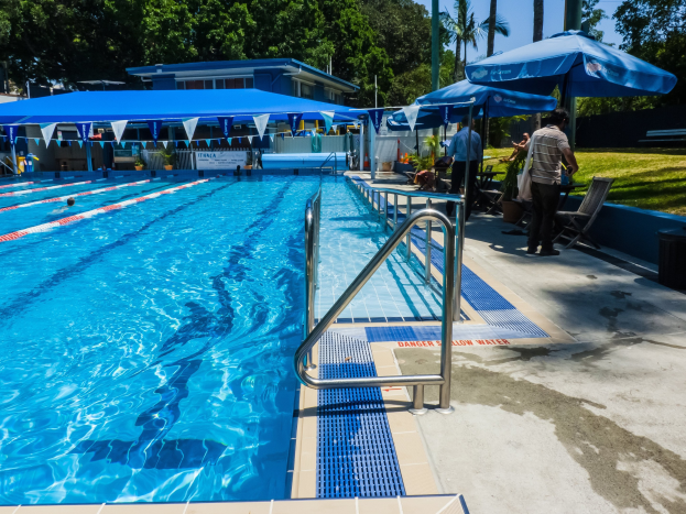 Großer rechteckiger Swimmingpool mit schwimmenden Menschen, umgeben von Geländern, Sonnenschirmen, Liegestühlen, Tischen, Grünpflanzen und einem Gebäude unter einem klaren blauen Himmel.