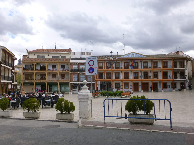 Ein belebter Stadtplatz mit Menschen auf Stühlen sitzend und stehend, umgeben von Topfpflanzen, Metallabsperrungen, Straßenlaternen mit Flaggen, ein Schild, Gebäude mit Fenstern und einem bewölkten Himmel.