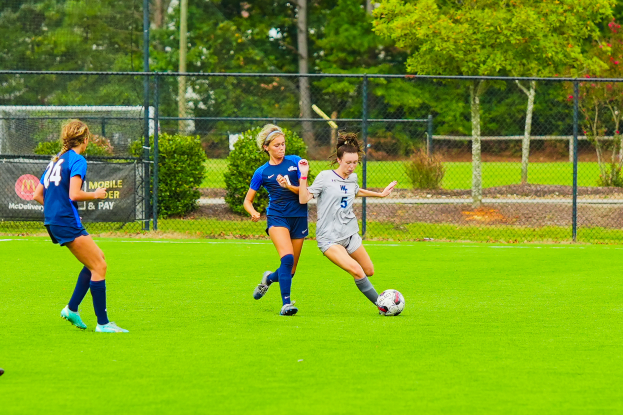 Eine Gruppe junger Frauen beim Fußballspielen auf einem Feld umgeben von Bäumen, Pflanzen, Maschendrahtzaun, Pfosten und einem Banner mit Text, mit einer Person im Hintergrund in Turnschuhen.