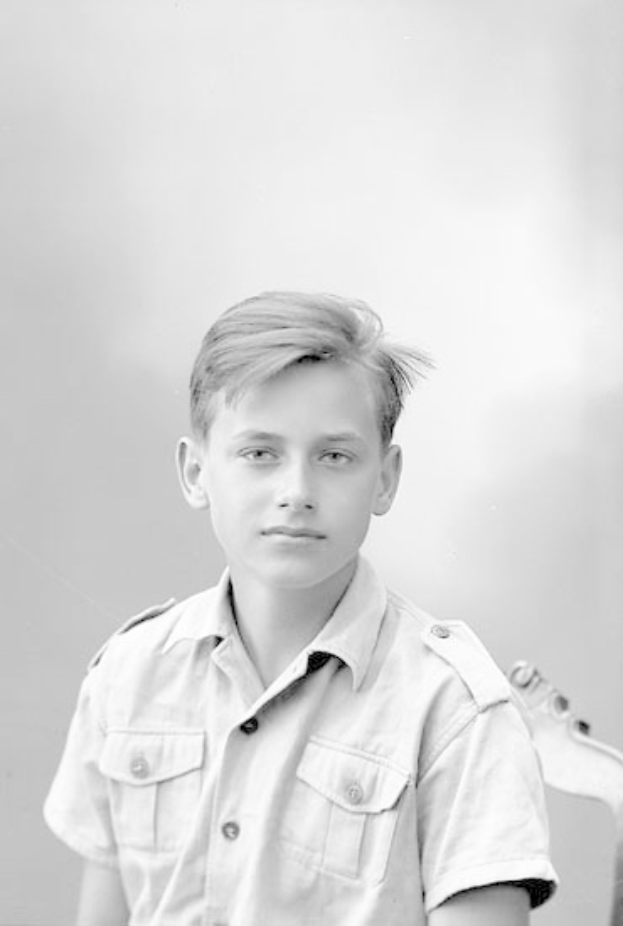 Ein junger Junge in einem Schwarz-Weiß-Foto hält eine Gitarre und trägt ein Hemd mit ernster Miene, kurzem dunklen Haar und konzentriertem Blick auf das Instrument.