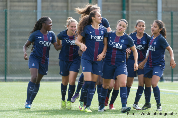 Eine Gruppe junger Frauen, die auf einem Rasenfußballfeld mit Maschendrahtzaun und einer Wand im Hintergrund spielen, und "Paris Saint-Germain Women's Soccer"-Text in der unteren rechten Ecke.