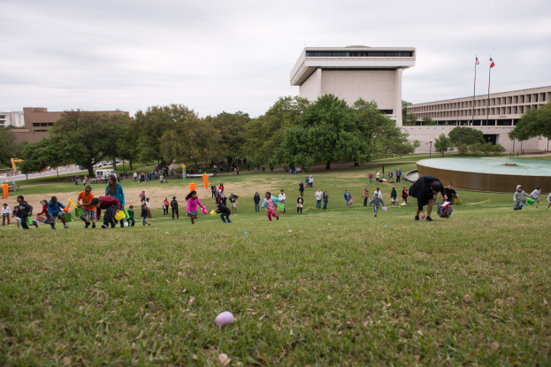 Gruppe von Menschen beim Ostereiersuchen auf einer grünen Wiese mit Bäumen, Gebäuden, Fahnen, einem Brunnen und einem bewölkten Himmel im Hintergrund.