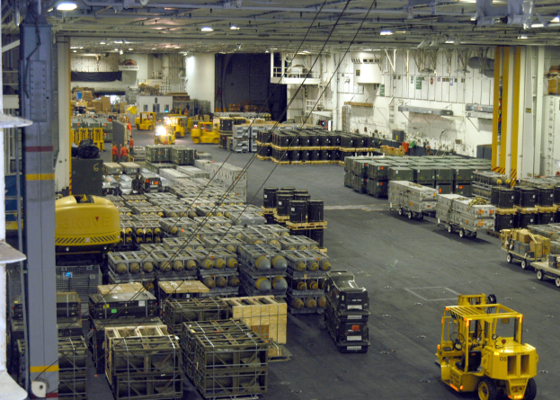 Ein großes Lagerinnere voller Lkw, Container, Menschen, Lampen, Stangen und anderen Gegenständen mit einem sichtbaren Rad.