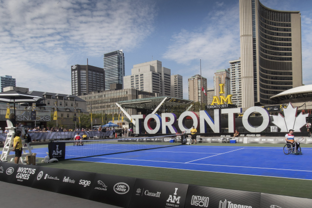 Eine Gruppe von Menschen, die Tennis auf einem Platz mit einer Stadtkulisse im Hintergrund spielt, umgeben von Zuschauern, Bannern, Gebäuden, Bäumen, Polen, Fahnen und einem bewölkten Himmel.