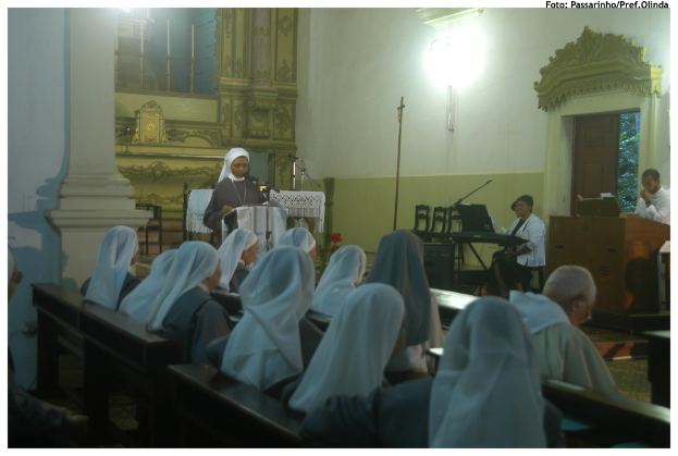 Eine Gruppe von Menschen auf einer Bank sitzend, eine Frau an einem Podium stehend, mit Stühlen, mehr Menschen an einem anderen Podium und einer Wand im Hintergrund.