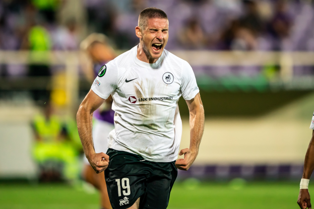 Ein Mann in weißem Hemd und schwarzen Shorts rennt auf einem Fußballfeld, feiert ein Tor, umgeben von einer Gruppe von Menschen, mit einem unscharfen Hintergrund.