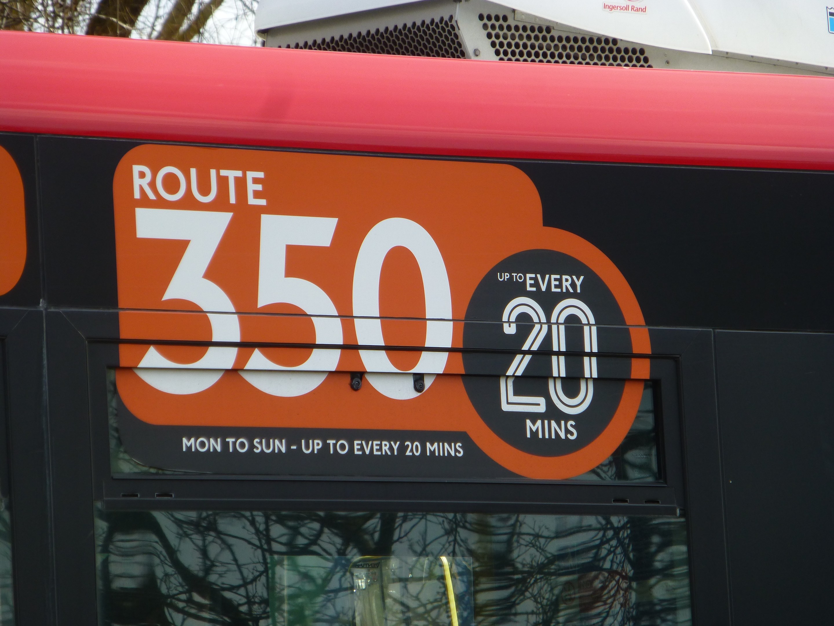 Roter Doppeldeckerbus mit der Aufschrift "Route 350", der durch einen baumbestandenen Bereich unter einem klaren Himmel fährt.