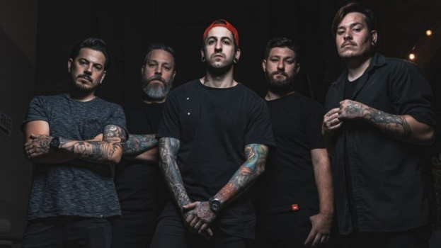 Eine Gruppe von Männern, die zusammen in einem schwach beleuchteten Raum stehen, von rechts beleuchtet, während sie offenbar ein Interview für Avenged Sevenfold geben.