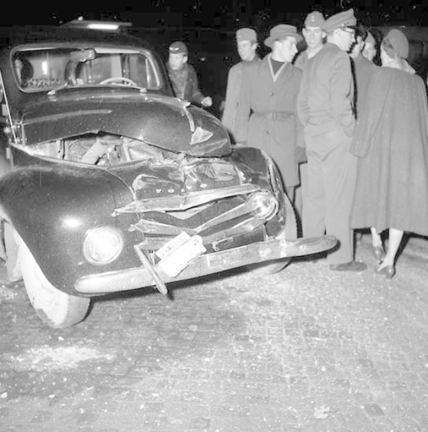 Schwarz-weiß-Foto eines schwer beschädigten Autos am Straßenrand mit eingedellter Motorhaube und zersplitterten Fenstern, umgeben von besorgten Menschen.