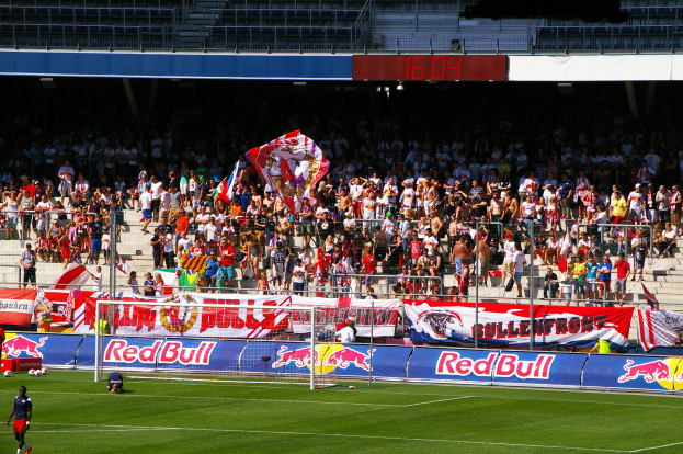 Ein Fußballspiel in einem Stadion mit Spielern auf dem Feld, ein Torpfosten mit Netz, Banner, ein Metallzaun, eine Anzeigetafel, ein Bildschirm und ein Dach mit Deckenbeleuchtung.