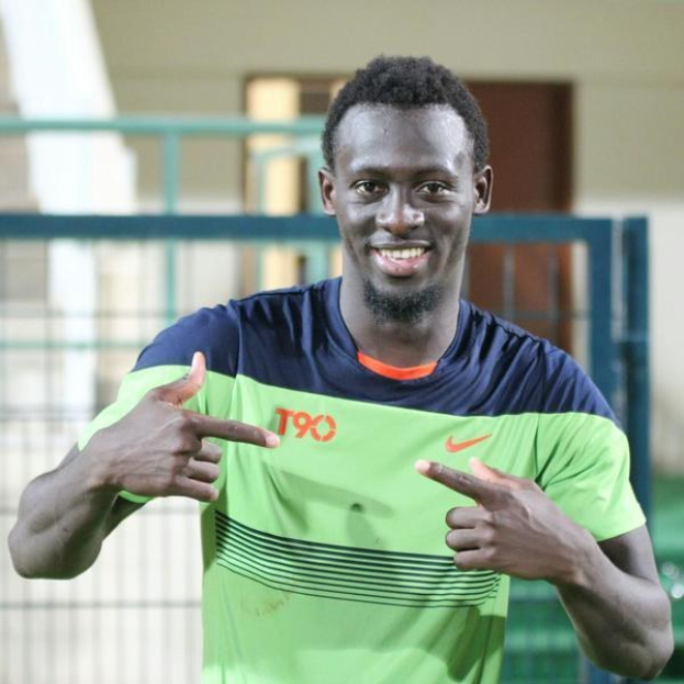 Ein Mann in einem grünen und blauen Hemd zeigt mit einem Lächeln auf die Kamera, während er vor einem unscharfen Geländer, einer Wand und einer Tür steht.