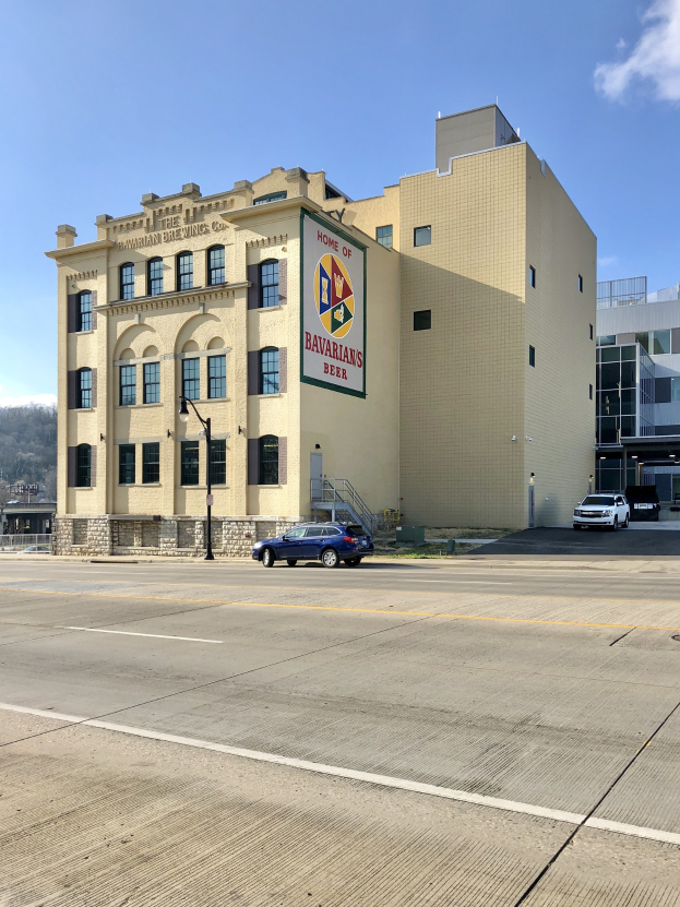 Ein großes Gebäude mit einer Tafel, auf der "Home of Bavarian Brewing Co." steht, umgeben von Straßenlaternen, Autos, Bäumen, Hügeln und einem bewölkten Himmel.