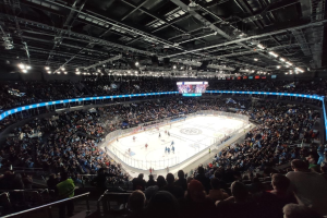Hockey-Spiel in einer großen Arena mit Zuschauern auf den Tribünen und einigen Menschen am Boden, mit Geländern, Überwachungsbildschirmen und heller Beleuchtung, identifiziert als O2 Arena in London.