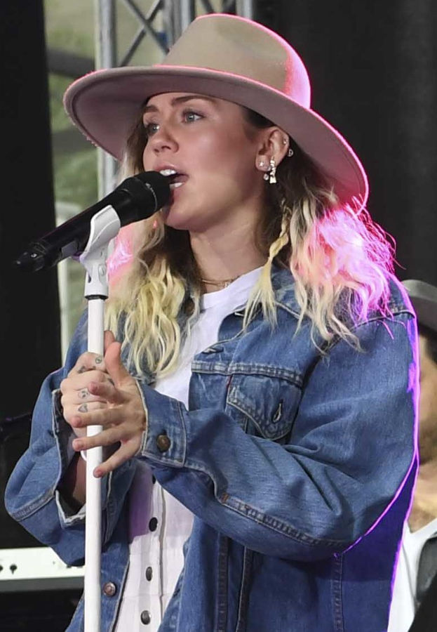 Miley Cyrus bei der CMA Music Festival in Nashville, Tennessee, singend in ein Mikrofon mit Ständer, während sie eine Mütze trägt, mit einer Person in einer Kappe rechts von ihr und sichtbaren Musikinstrumenten im Hintergrund.