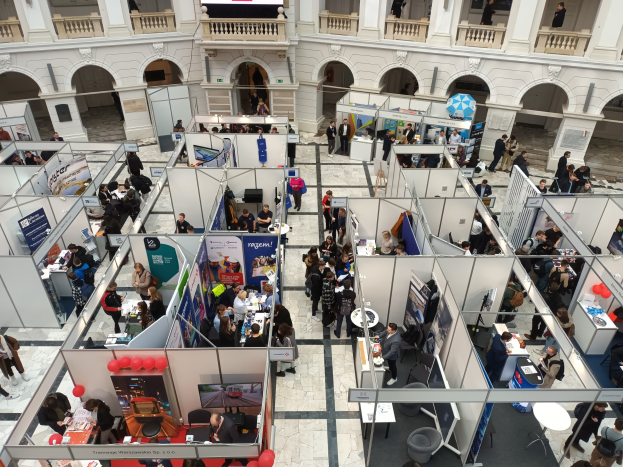 Eine große Gruppe von Menschen auf einer internationalen Fachmesse mit Ständen, Bannern, Tischen, Stühlen und Ballons in einem Raum mit architektonischen Bögen, Säulen und einem Hintergrundbild.