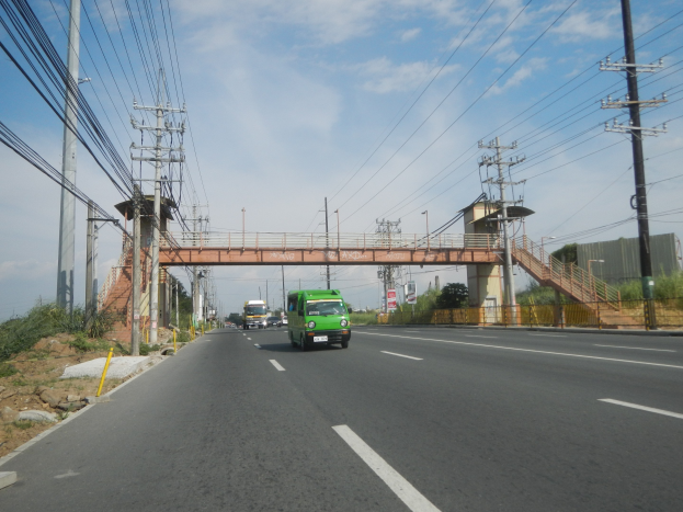 Grüner Lkw fährt auf einer Straße neben einer Brücke mit elektrischen Masten und Drähten entlang der Straße, Bäume und Gebäude im Hintergrund unter einem klaren blauen Himmel.