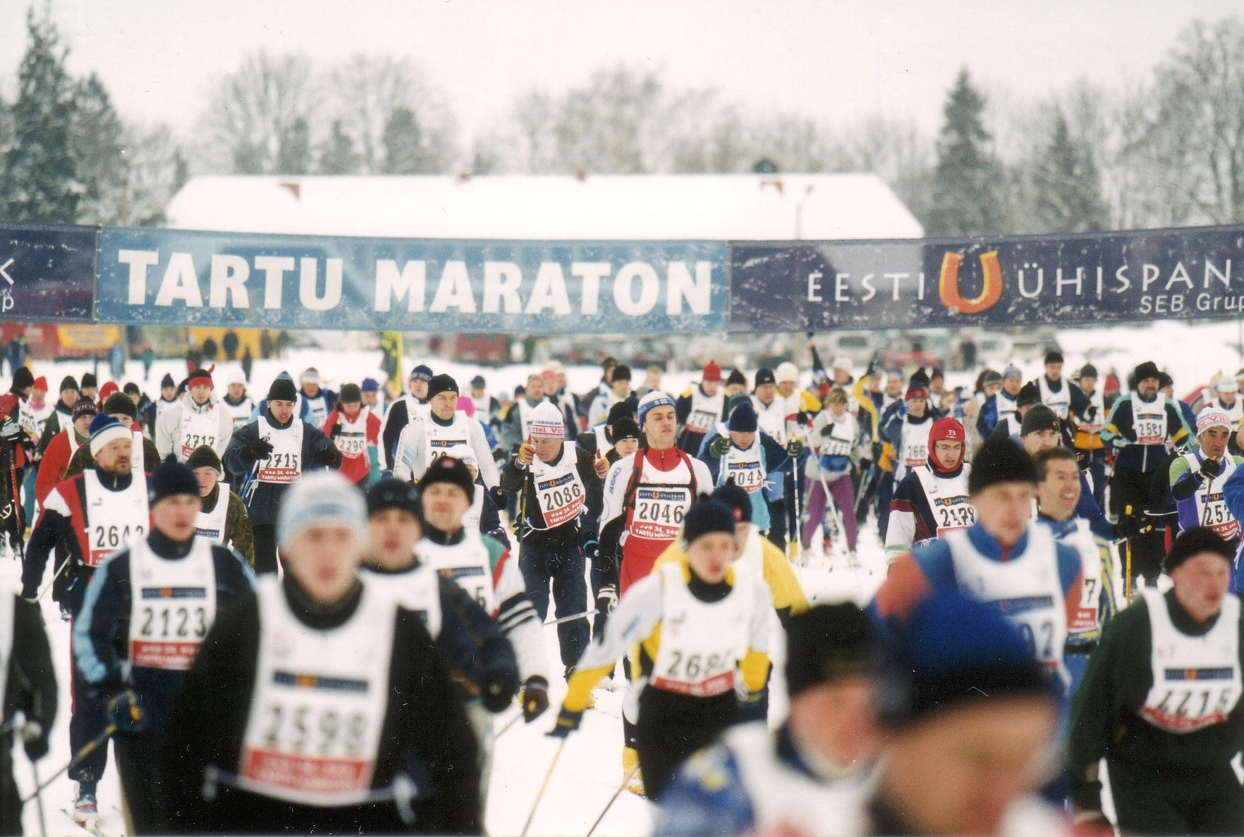 Gruppe von Menschen, die auf Skiern laufen, mit Mützen und Ski-Stöcken, sowie Bannern, Bäumen und einem klaren blauen Himmel im Hintergrund; Text unten.