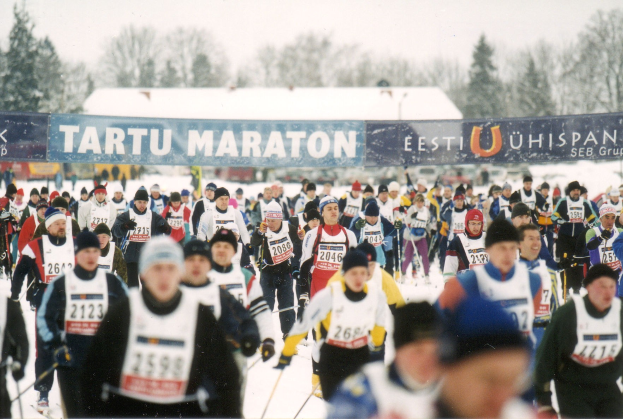 Gruppe von Menschen, die auf Skiern laufen, mit Mützen und Ski-Stöcken, sowie Bannern, Bäumen und einem klaren blauen Himmel im Hintergrund; Text unten.