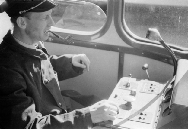 Schwarz-weißes Bild eines Mannes in Mütze und Uniform, der auf den Kontrollen eines Zugs sitzt und eine Pfeife raucht, mit Glasfenstern im Hintergrund.