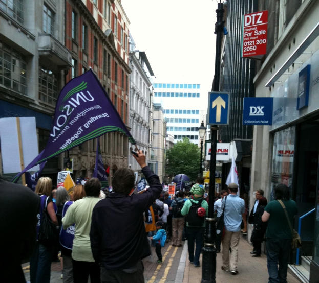Eine große Gruppe von Menschen auf einer Straße, einige halten Fahnen und Schilder, mit einem Schildständer, Gebäuden mit Fenstern, einem Baum, einem Lichtmast und einem Himmel im Hintergrund.