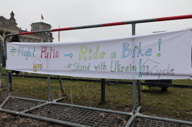 Eine Gruppe von Menschen hält ein Banner mit der Aufschrift "Fight Putin, Ride a Bike, Stand with Ukrainian U-Cycle" und steht auf dem Boden neben einem Metallzaun, trockenen Blättern, einem Gebäude mit Fenstern, einem Fahnenmast mit einer Fahne und einem bewölkten Himmel.