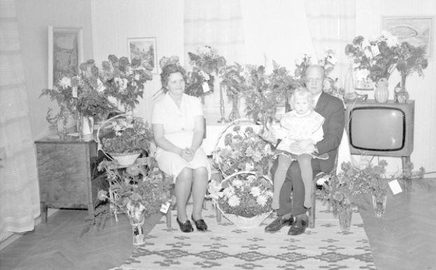 Schwarz-weiß-Foto einer Familie aus drei Personen - einem Mann, einer Frau und einem Kind - auf einem Sofa in einem Wohnzimmer mit Blumenvasen, einem Teppich, Vorhängen und Fotorahmen im Hintergrund.