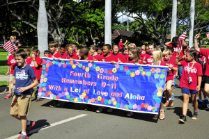 Gruppe von Kindern in roten Hemden, die eine Straße entlanggehen und eine Fahne sowie Flaggen halten, mit grasgesäumten Seiten, Bäumen, Pfählen, Häusern und Himmel im Hintergrund.