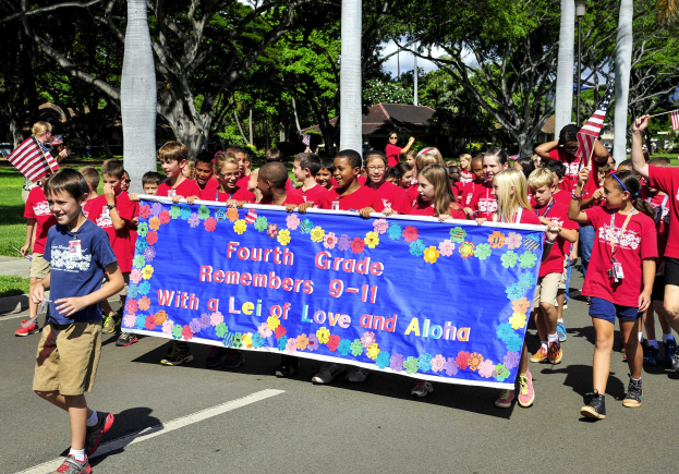Gruppe von Kindern in roten Hemden, die eine Straße entlanggehen und eine Fahne sowie Flaggen halten, mit grasgesäumten Seiten, Bäumen, Pfählen, Häusern und Himmel im Hintergrund.