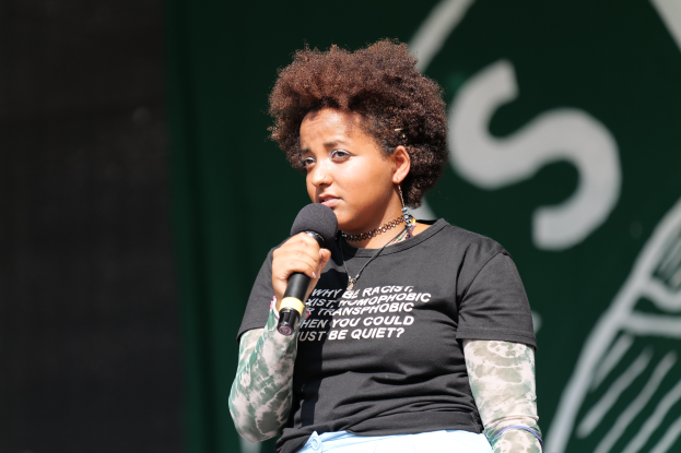 Eine Frau steht selbstbewusst auf einer Bühne, hält ein Mikrofon in der Hand und trägt ein schwarzes T-Shirt und Jeans, ihre Haare sind zu einem Pferdeschwanz gebunden.