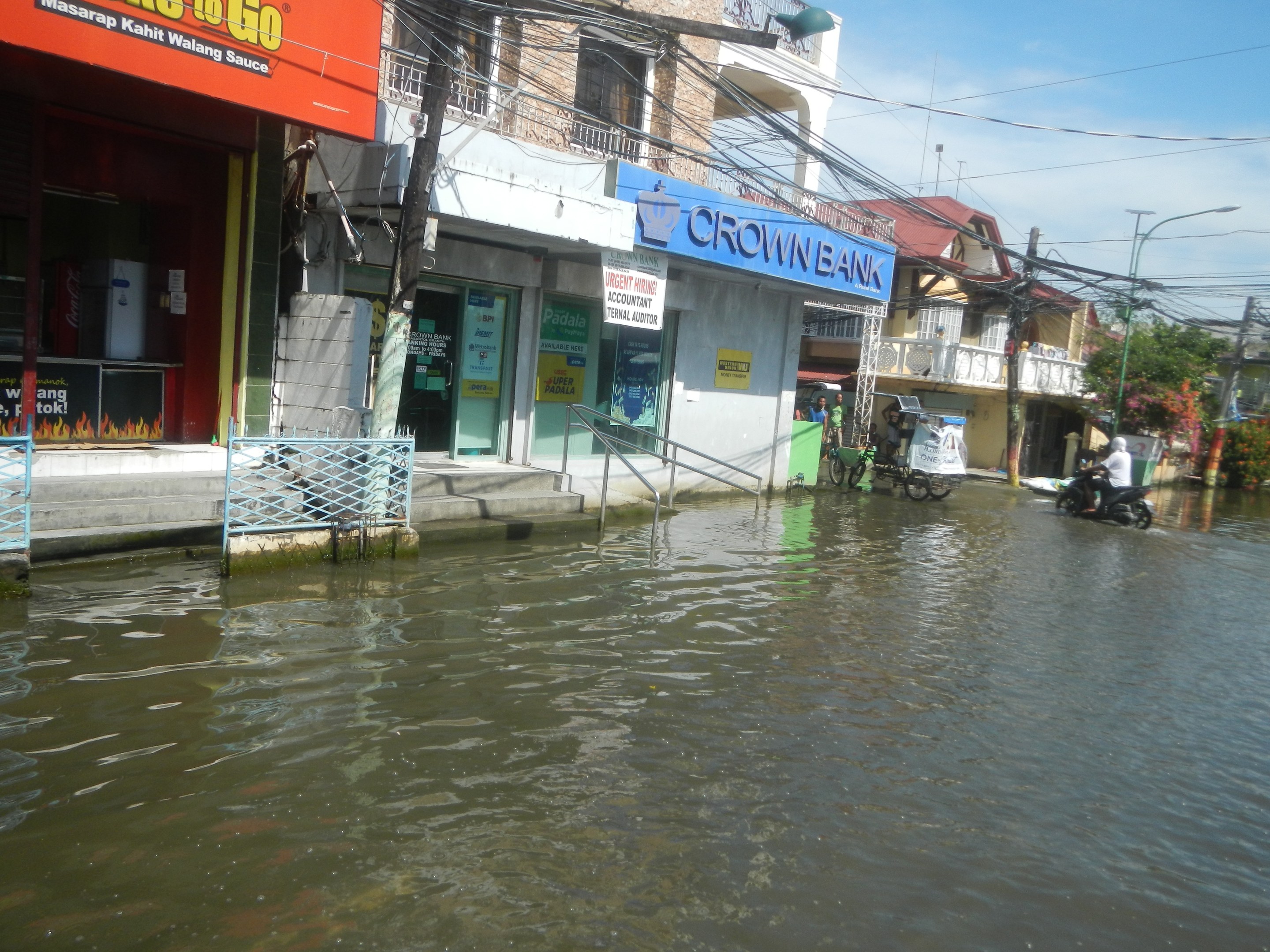 Überschwemmte Stadtstraße mit Gebäuden, Fahrzeugen, Fußgängern und Infrastruktur teilweise unter Wasser.