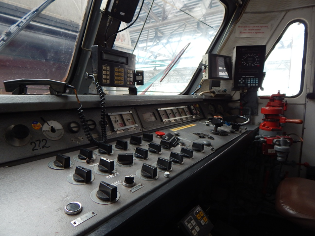 Eine Zugsteuerkonsole mit Knöpfen, Schaltern und Messgeräten im Vordergrund und Fenstern an den Seiten, mit einem Zug im Hintergrund.