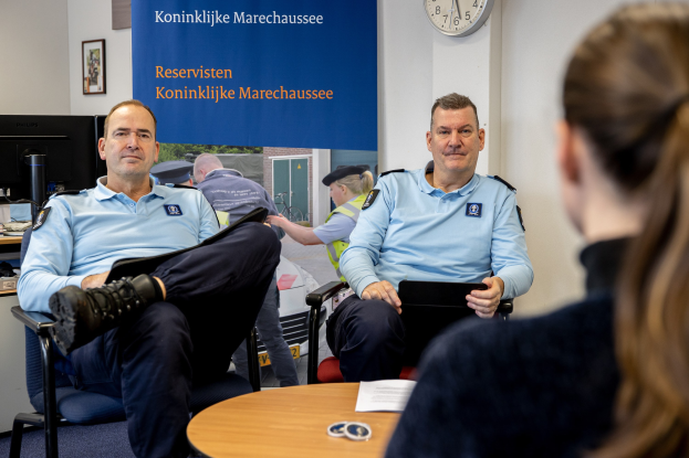 Zwei Polizeibeamte sitzen an einem Tisch mit einer Frau, Papieren und Gegenständen auf dem Tisch, einem Wandbanner und einer Uhr im Hintergrund, einem zweiten Tisch mit einem Monitor, Tastatur, Maus und Gegenständen links daneben und Fotorahmen an der Wand.