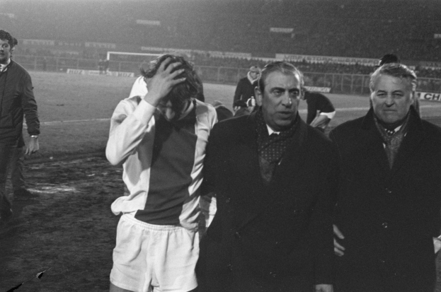 Schwarzes und weißes Bild einer Gruppe von Männern, die auf einem Fußballfeld stehen und ein Stadion im Hintergrund haben, die enttäuscht nach einem Sieg aussehen.