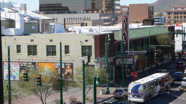 Eine belebte Stra├če mit verschiedenen Fahrzeugen, darunter ein Bus, Autos und ein Landfahrzeug, umgeben von Pfosten, Lichtern, Schildern, B├Ąumen, Geb├Ąuden, einem H├╝gel und einem klaren Himmel.